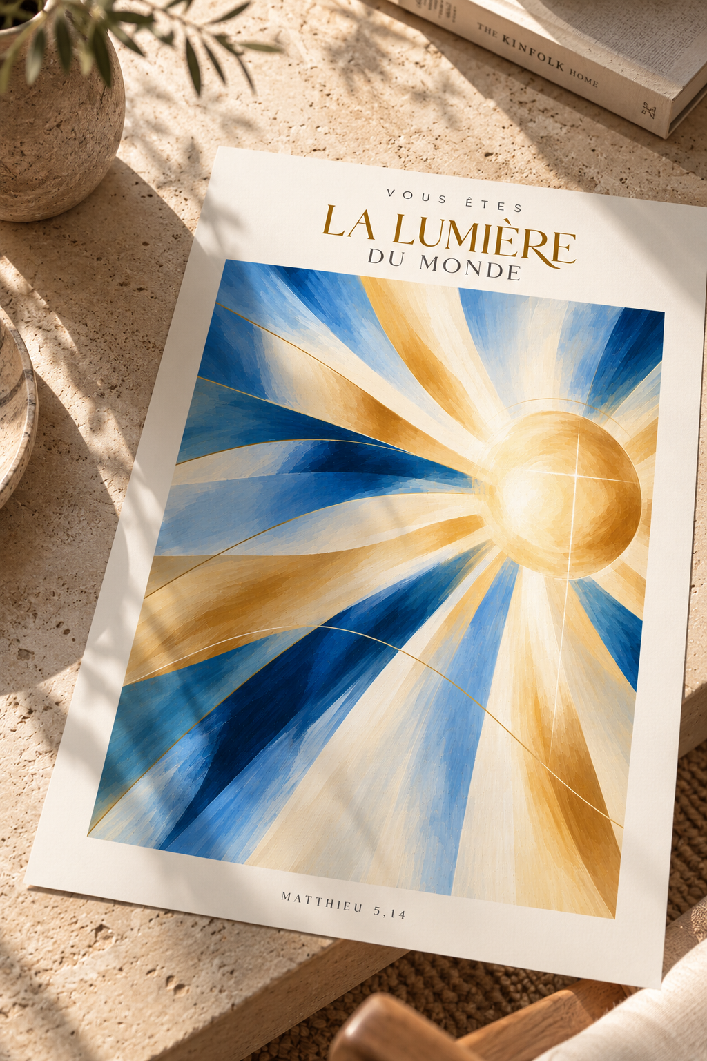 A printed art card or poster titled 'VOUS ÊTES LA LUMIÈRE DU MONDE' with a radiant sun illustration in blue, gold, and white, styled on a stone surface with a book and plant nearby.